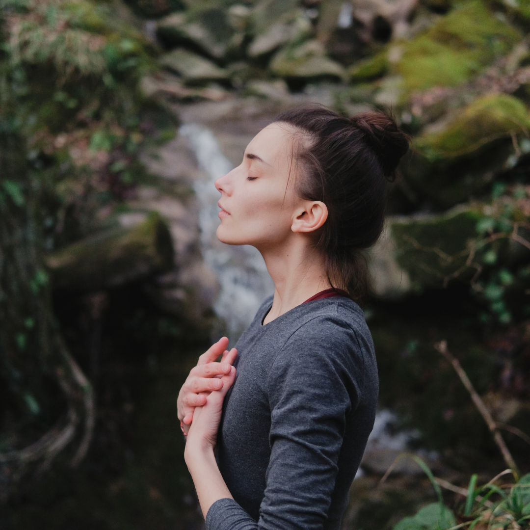 Frau vor Wasserfall mit Händen auf der Brust atmet tief durch